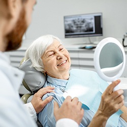 An older woman looking at her new smile in the mirror while her dentist explains the process of maintaining her implants at home